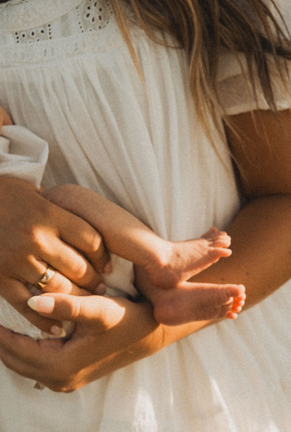 A mother in a white dress tenderly holds her newborn baby's tiny feet in her hands, wearing a gold ring.