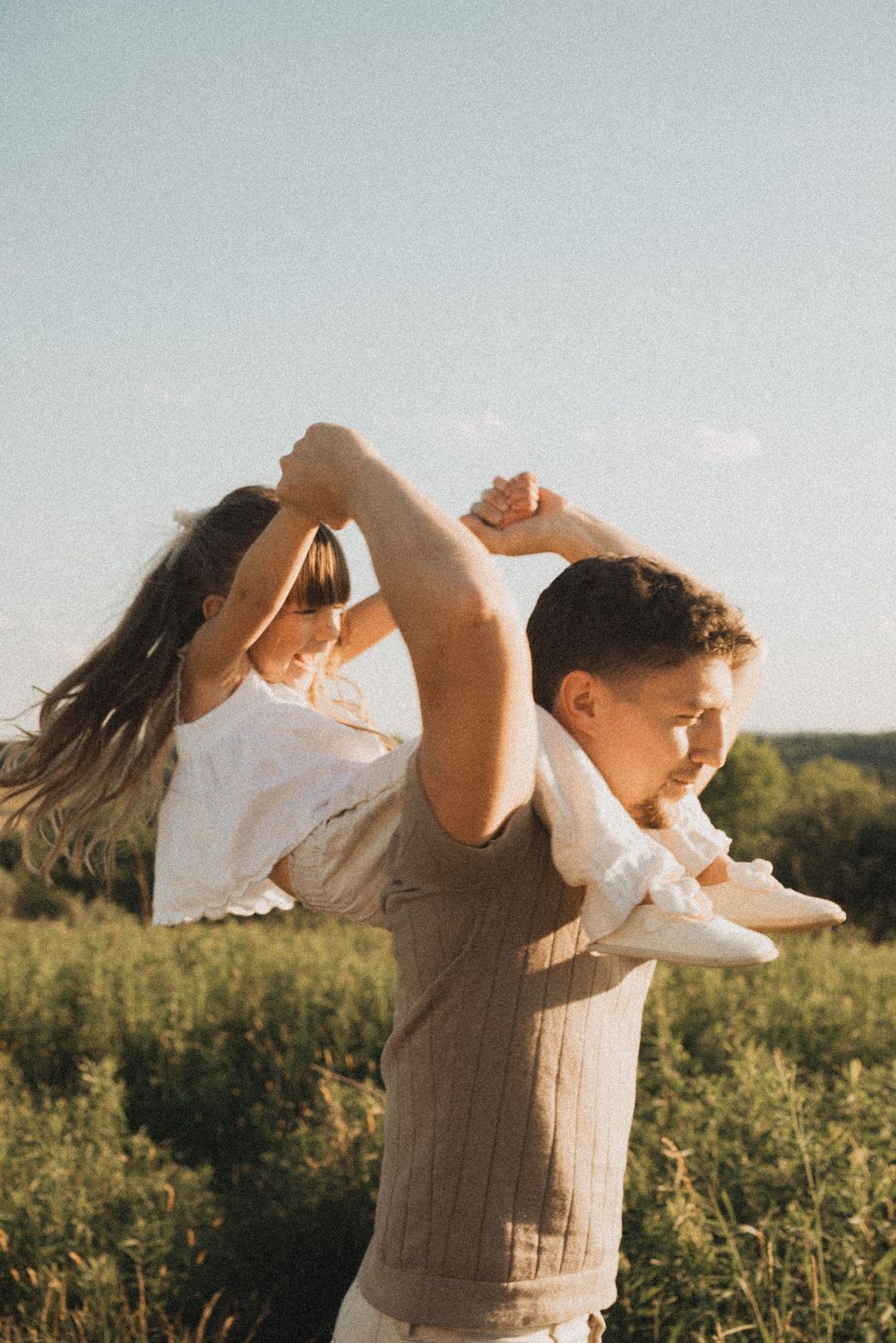 A man carries a young girl on his shoulders in a sunny green field, holding her hands as her hair flows in the breeze.