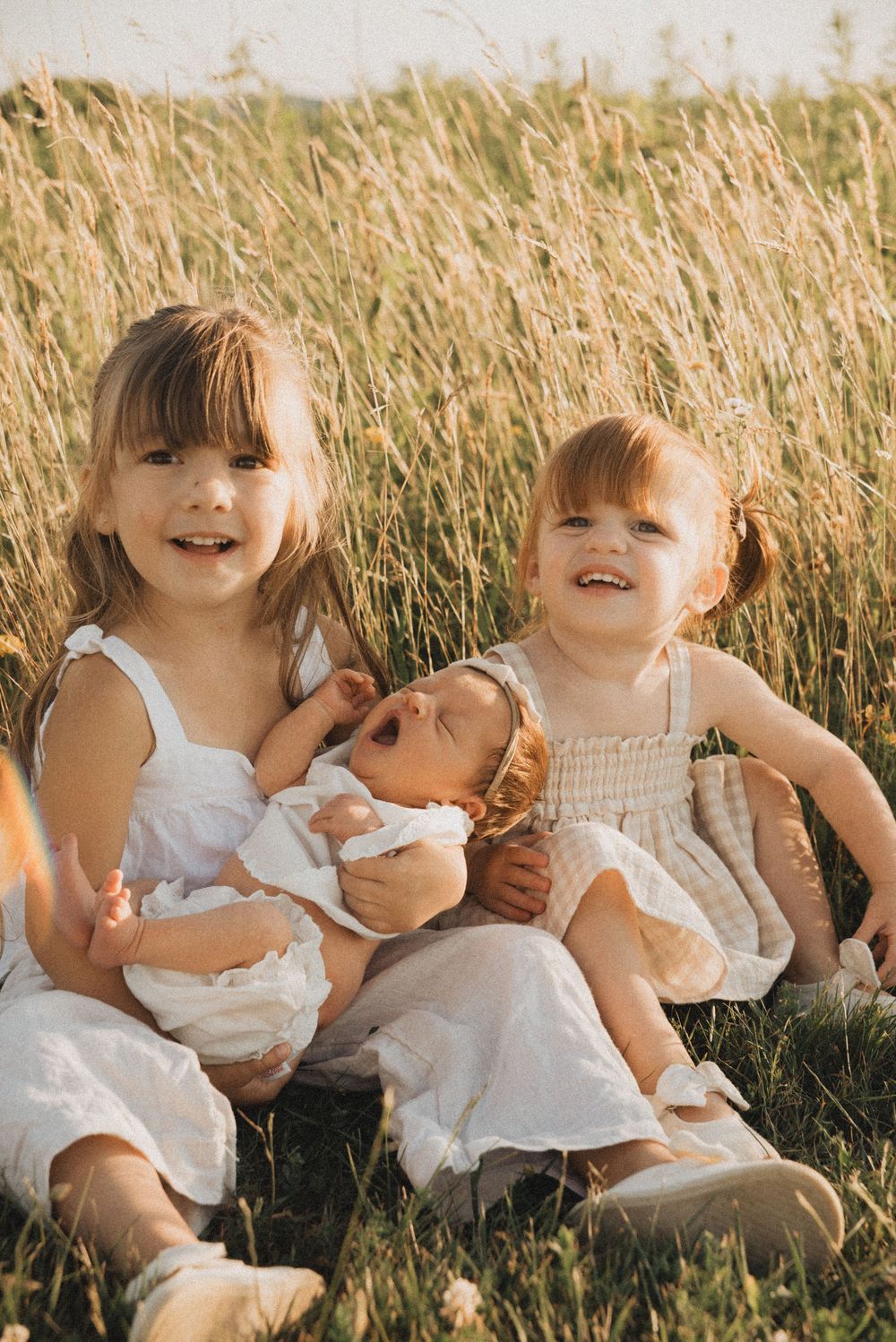 Two young girls sitting in a golden field, smiling and holding a newborn baby in warm sunlight.