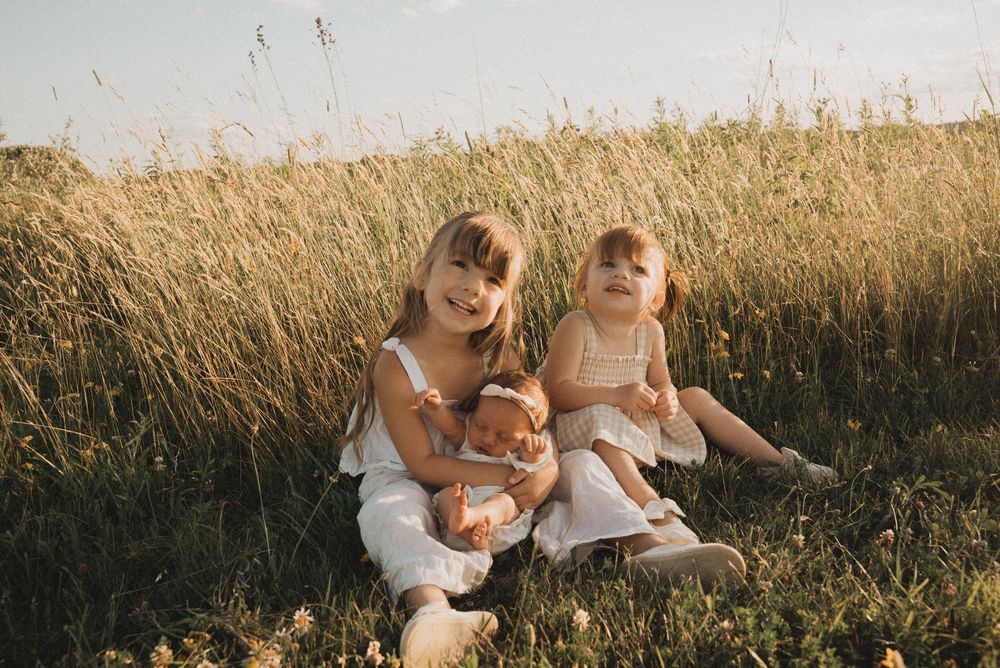 Two young girls in white dresses sitting in a sunny meadow, smiling, one holding a doll.