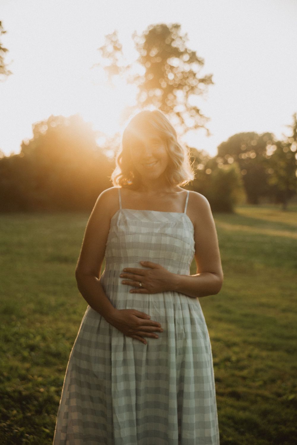Pregnant woman in plaid sundress holding baby bump at golden hour sunset in lush green field.