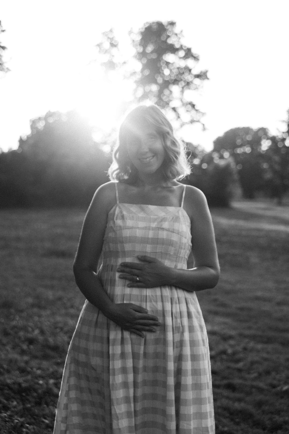 Black and white photo of a smiling pregnant woman in a plaid sundress cradling her baby bump outdoors at sunset.