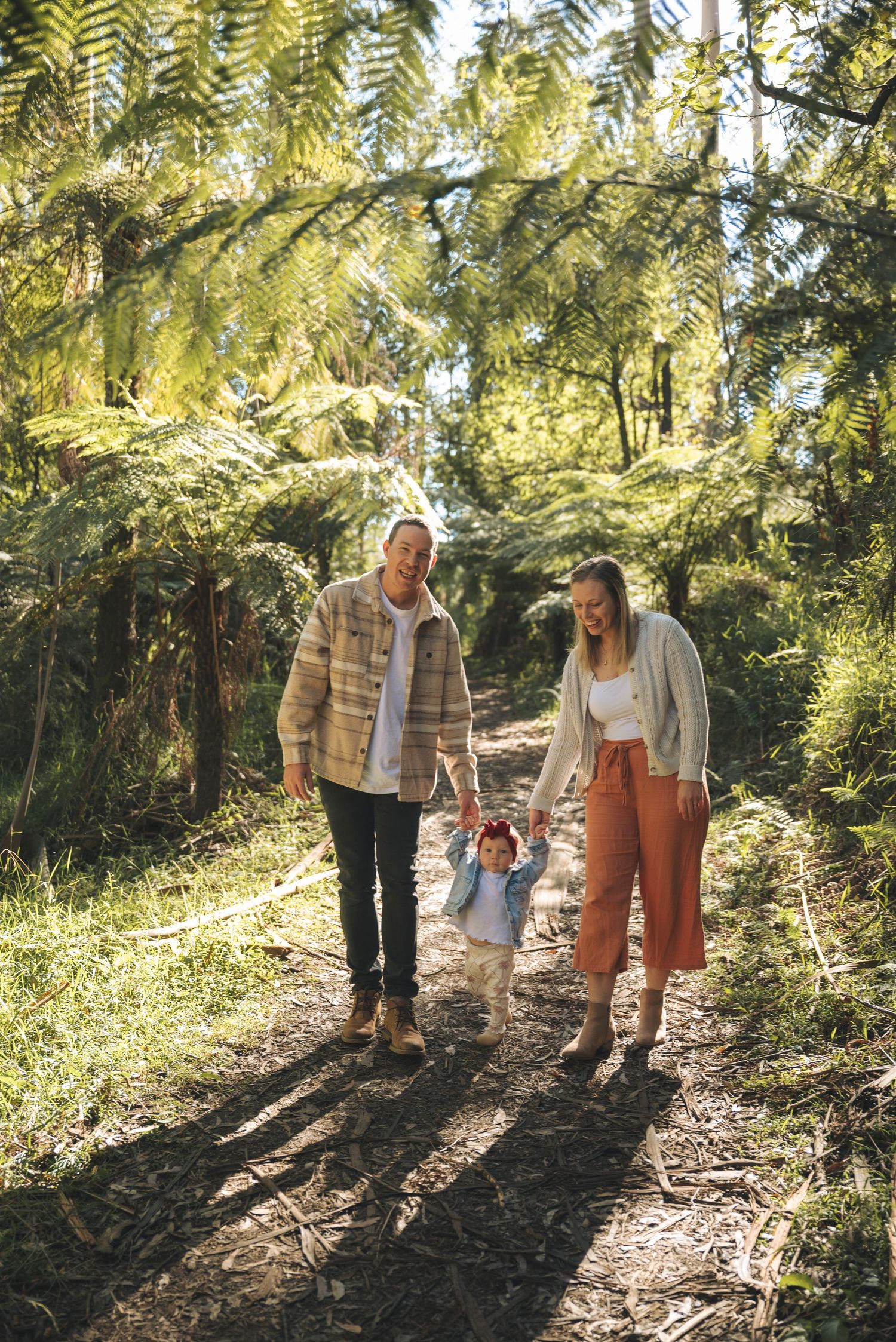 A young family walks hand-in-hand with their toddler along a sunlit forest path surrounded by lush green ferns.