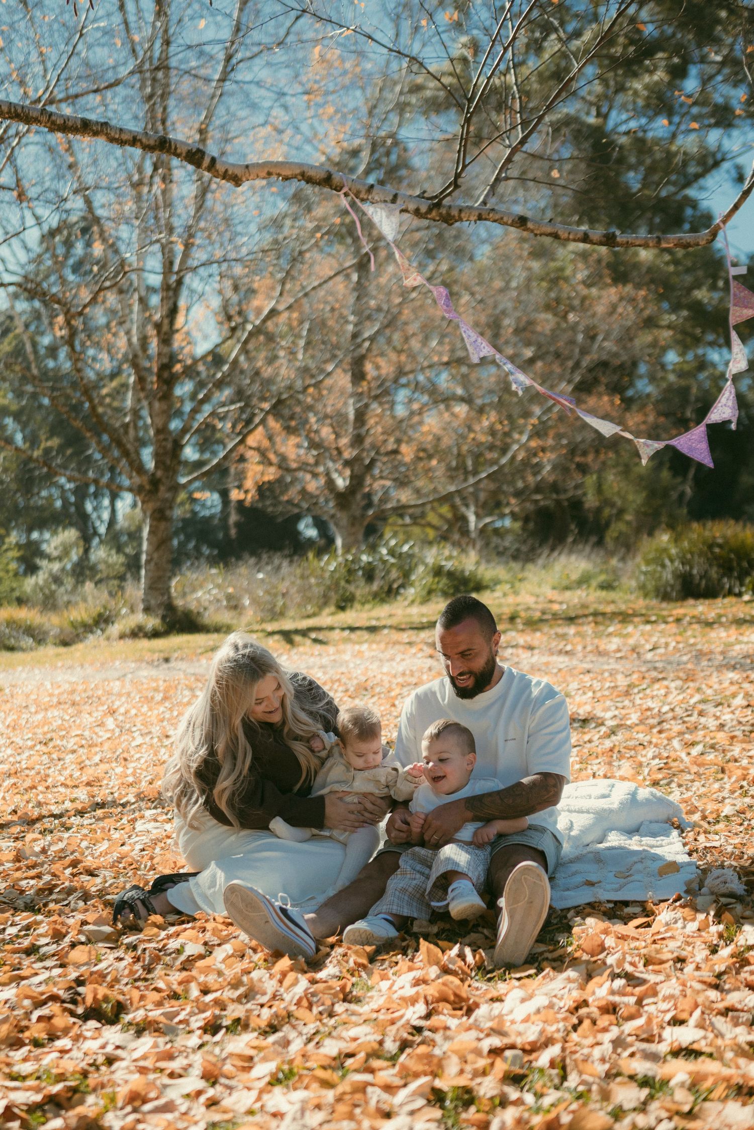 Family of four sitting on autumn leaves in a sunny park, with bunting decorations hanging from a bare tree branch.