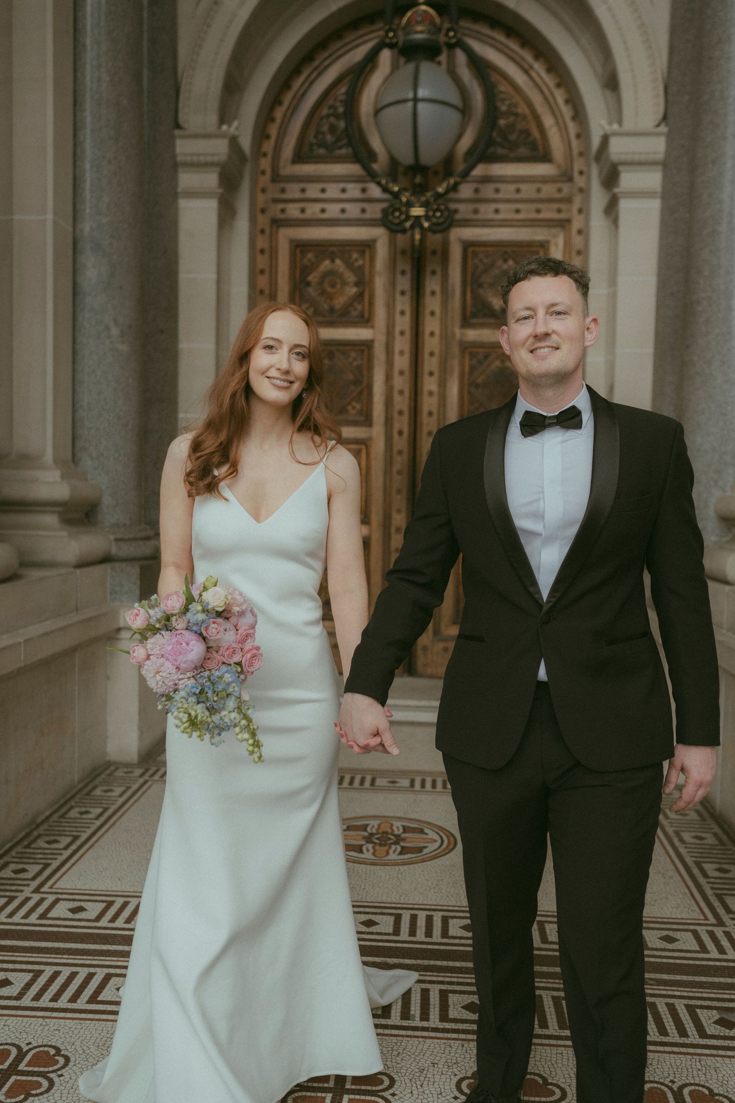 Bride and groom posing in front of ornate wooden door at elegant neoclassical building with decorative tile floor.