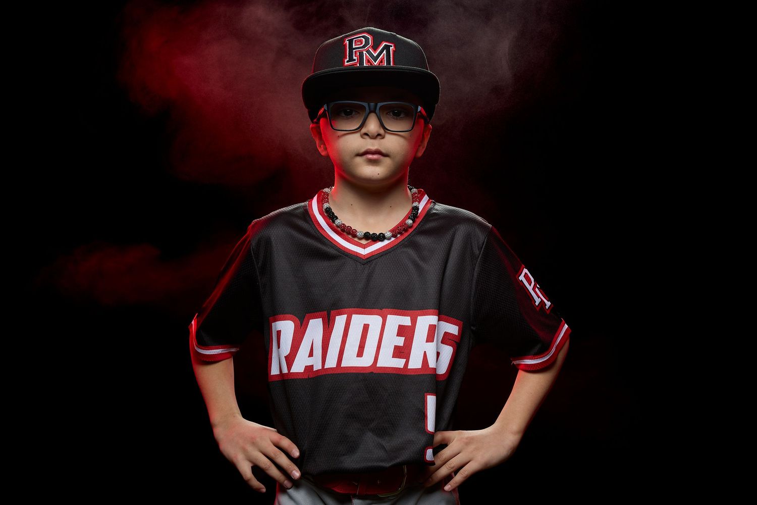 Young baseball player in black and red Raiders jersey and cap with glasses poses confidently on dark background.