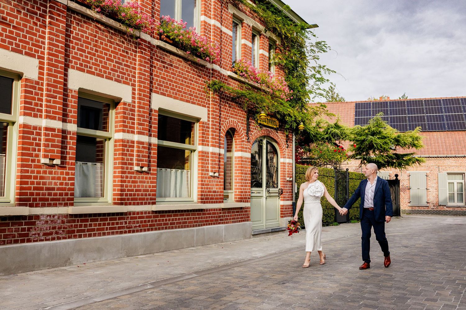 Bride and groom walking hand in hand outside a red brick building with solar panels on a sunny wedding day.