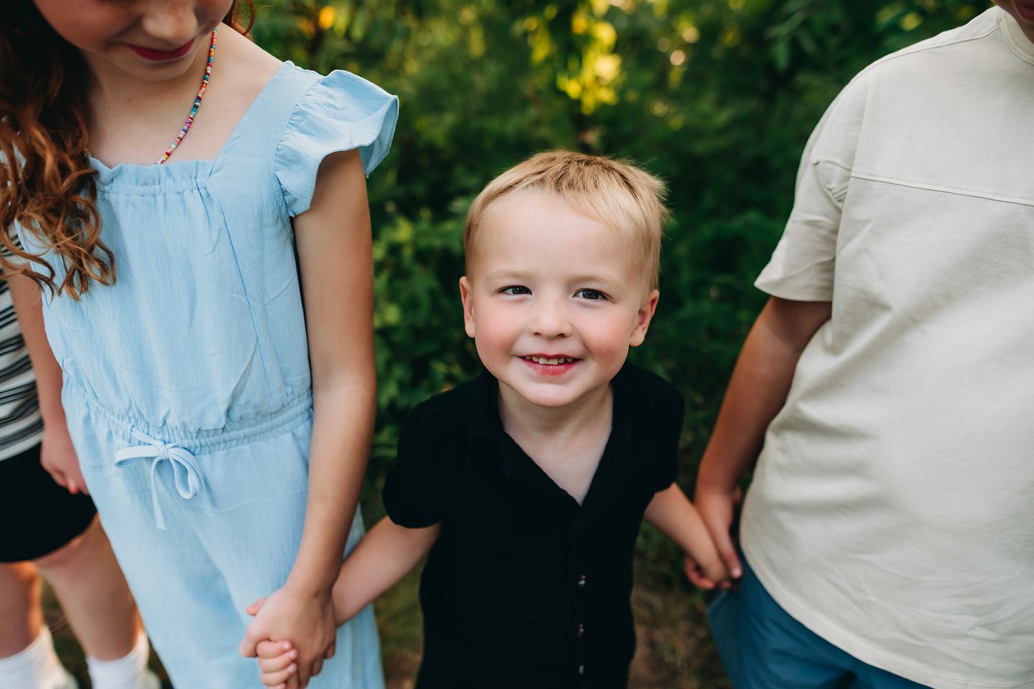 A smiling toddler boy in a black outfit holds hands with two older children outdoors among green trees.