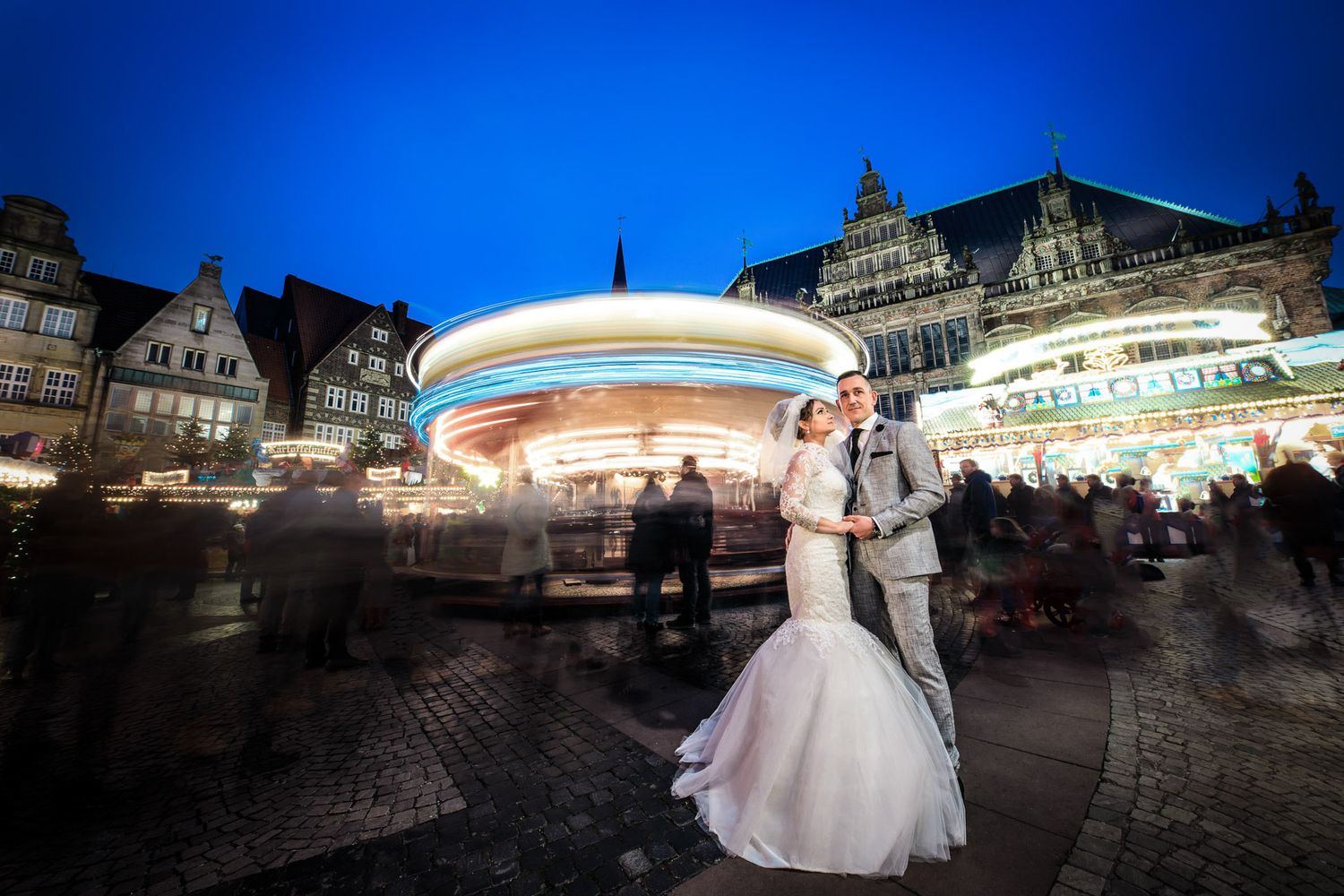 Bride and groom embrace in front of a glowing carousel at dusk in a European town square.
