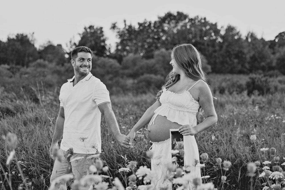 Black and white photo of a smiling couple holding hands in a flower field, woman cradling pregnant belly outdoors.