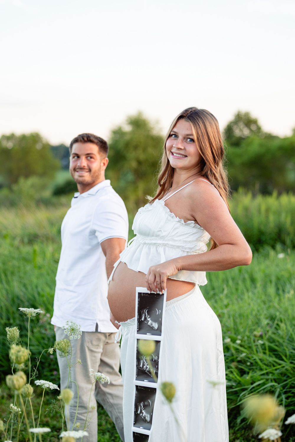 Pregnant woman in white dress holds ultrasound photos outdoors with smiling partner in green field at sunset.