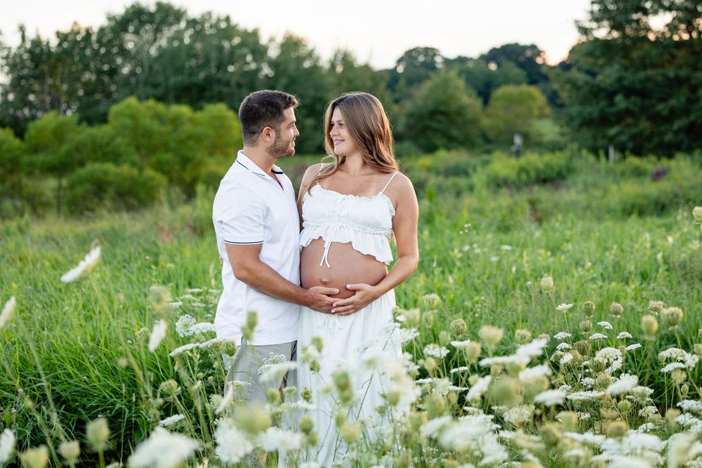 Couple in white outfits embracing in a flower field during a maternity photo shoot outdoors.