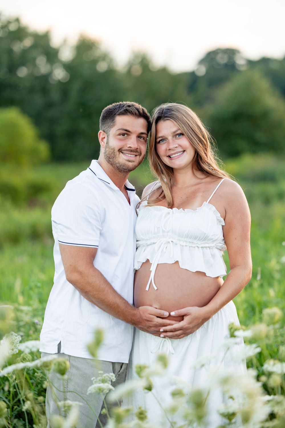 Smiling couple in white outfits outdoors, man embracing pregnant woman while both cradle her baby bump in a green field.