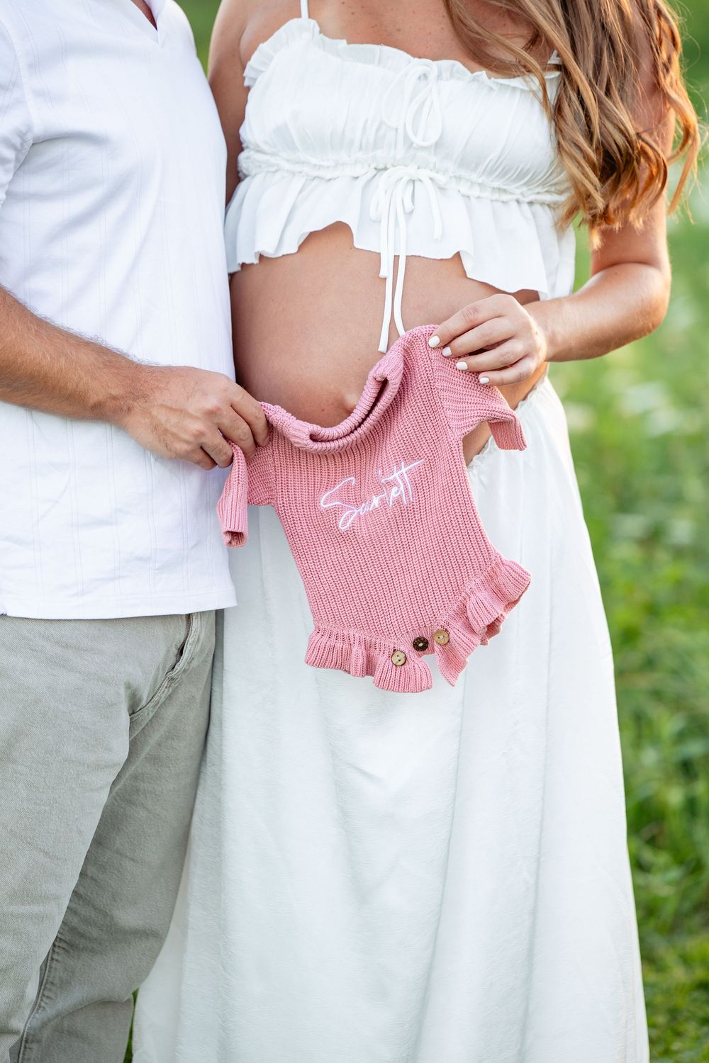 Couple holding pink knit baby onesie against pregnant belly outdoors.