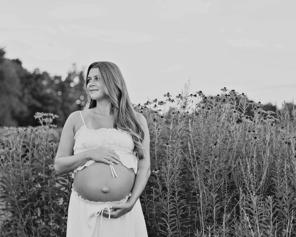 Black and white maternity photo of a pregnant woman in a white dress cradling her belly in a wildflower field.