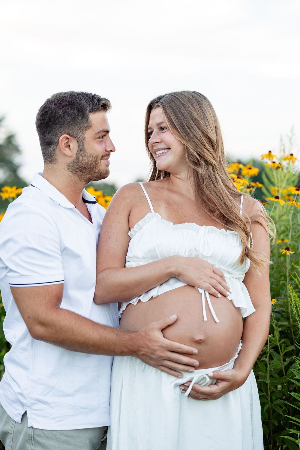 Happy pregnant couple in white embracing outdoors among yellow flowers, smiling at each other.