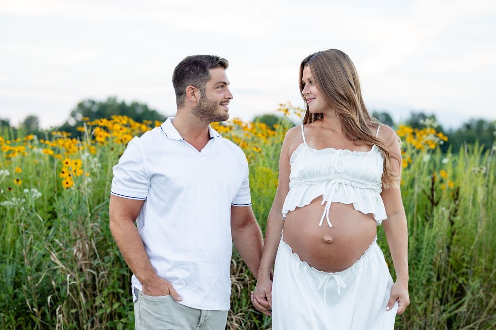Couple in white outfits holding hands in a flower field, woman showing pregnant belly, maternity photo shoot outdoors.