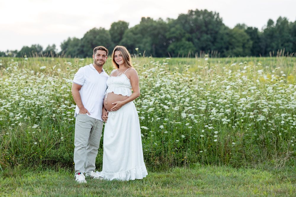 Pregnant couple posing in a white flower field, woman in white dress cradling baby bump, man in white shirt beside her.