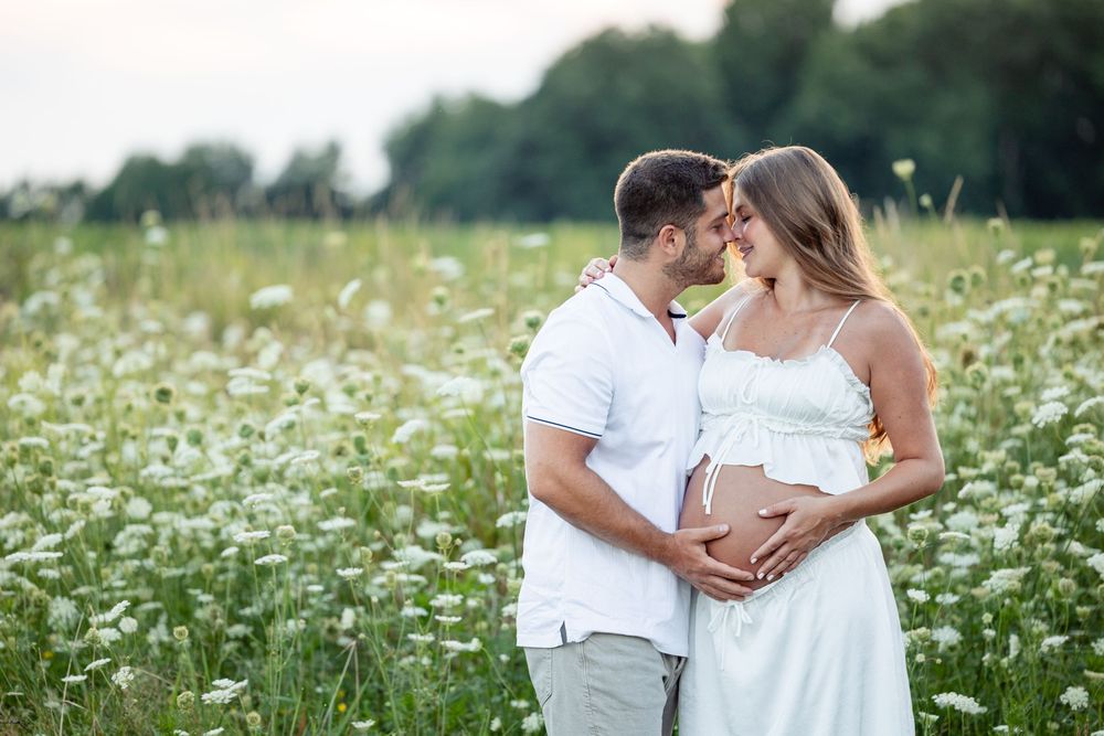 Expecting couple sharing a tender moment in a wildflower meadow, both dressed in white while cradling the pregnant belly.
