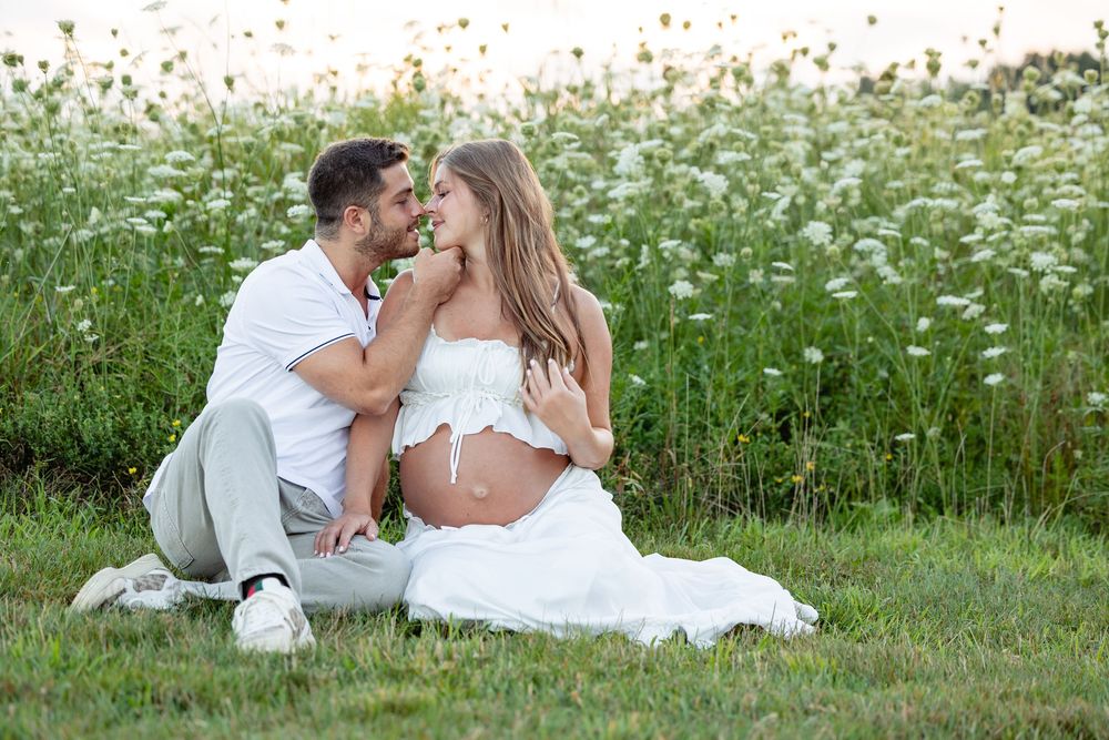 Couple sitting in a flower field, man kissing pregnant woman's cheek, both wearing white outfits.
