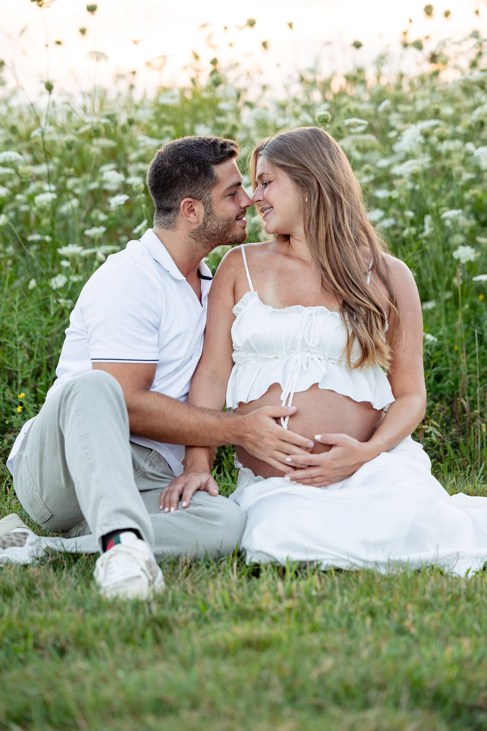 Pregnant couple sitting in a flower field, smiling at each other while man gently holds womans baby bump.
