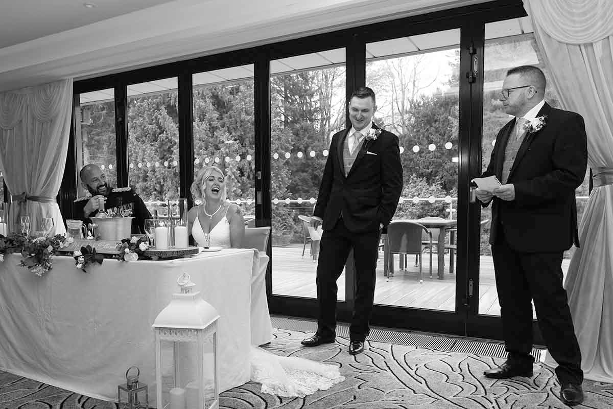 Black and white wedding photo of groom standing with best man giving a speech at elegant venue reception.