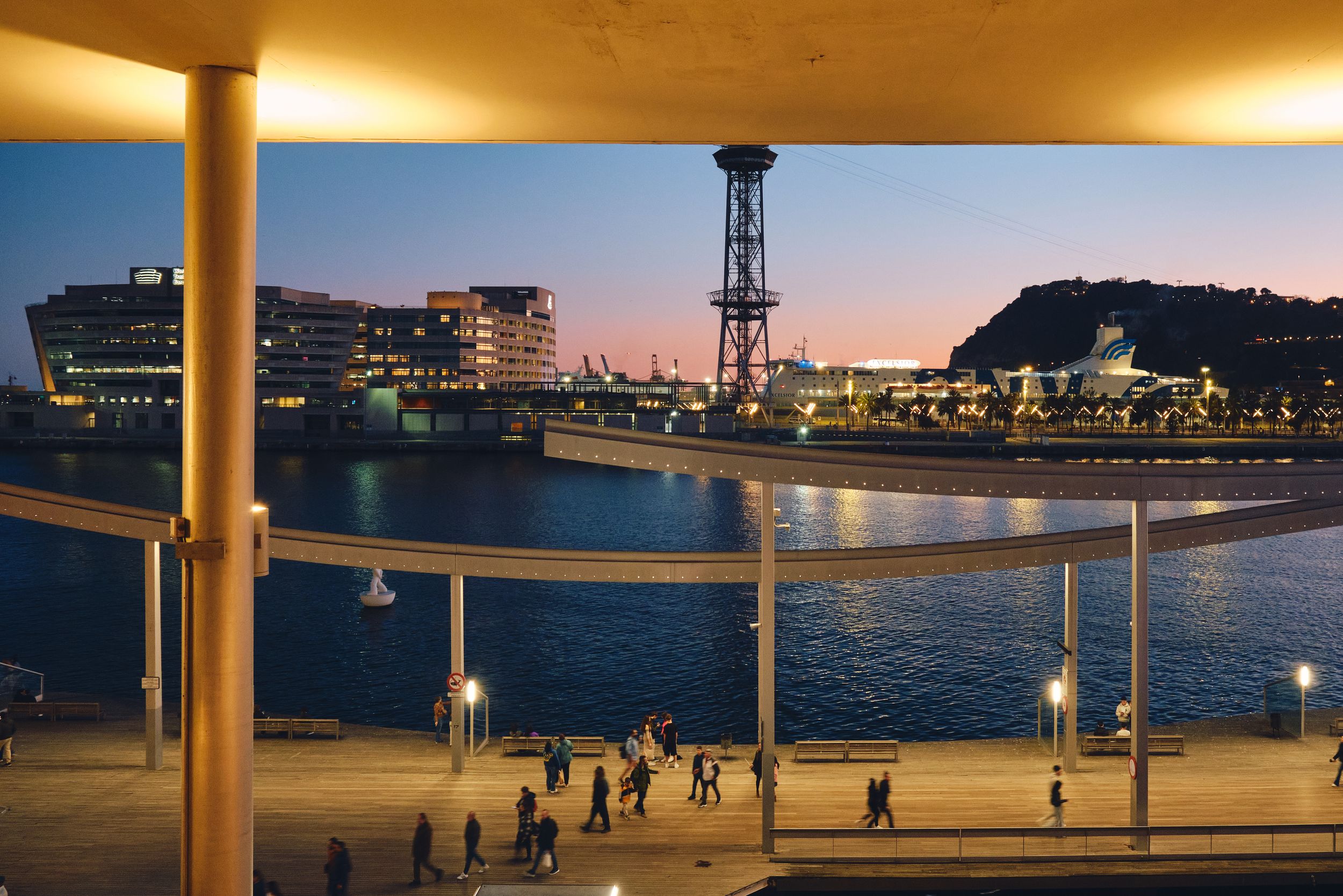 Twilight view of Barcelona harbor with illuminated waterfront promenade, cable car tower, and city skyline reflecting on calm water.