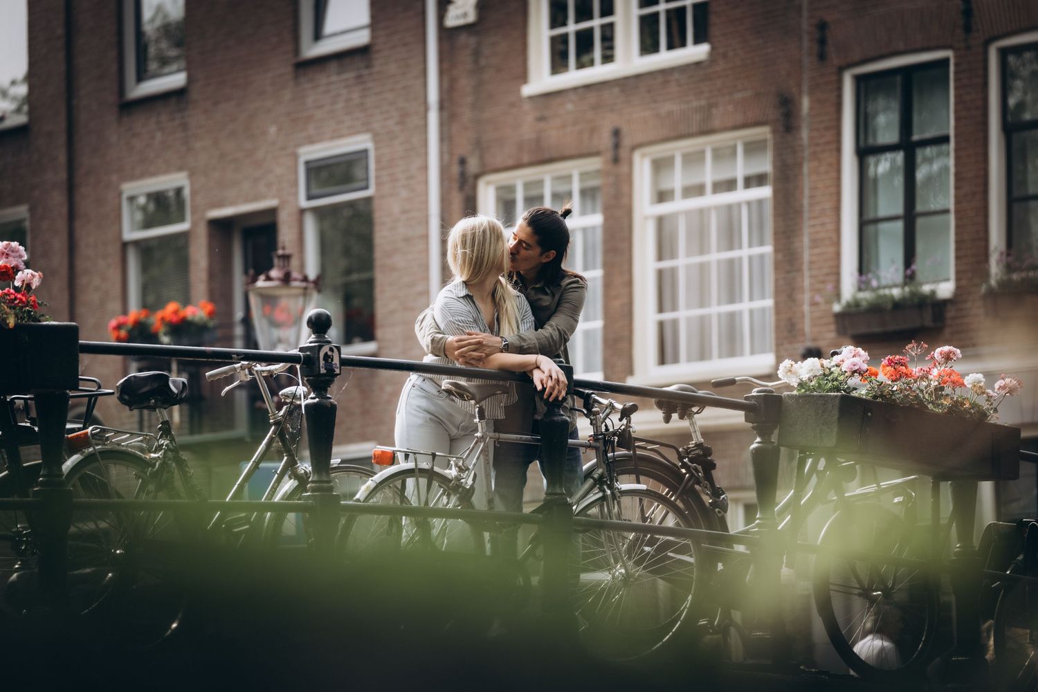 Paar steht auf einer Brücke in Amsterdam, er hält sie im Arm und küsst sie – Fahrräder im Hintergrund.
