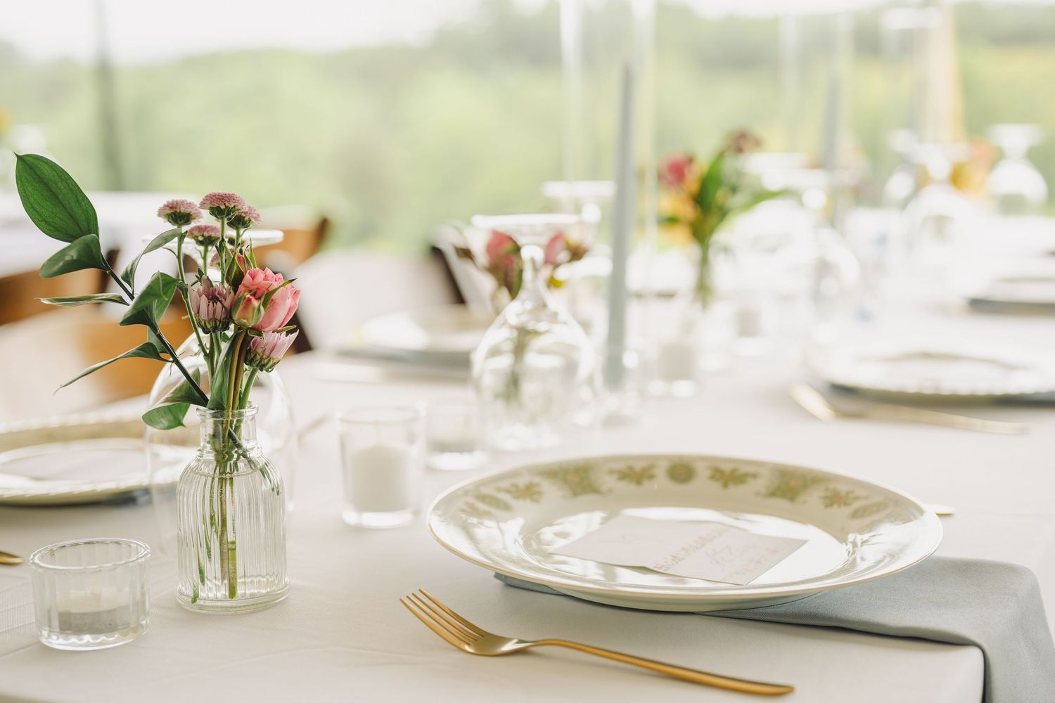 Elegant wedding table setting with gold cutlery, white plates, and pink floral centerpieces in glass vases.
