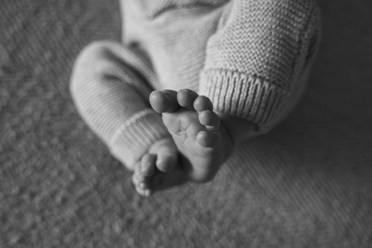 Black and white close-up of a newborn baby's tiny feet and toes in a knit onesie.