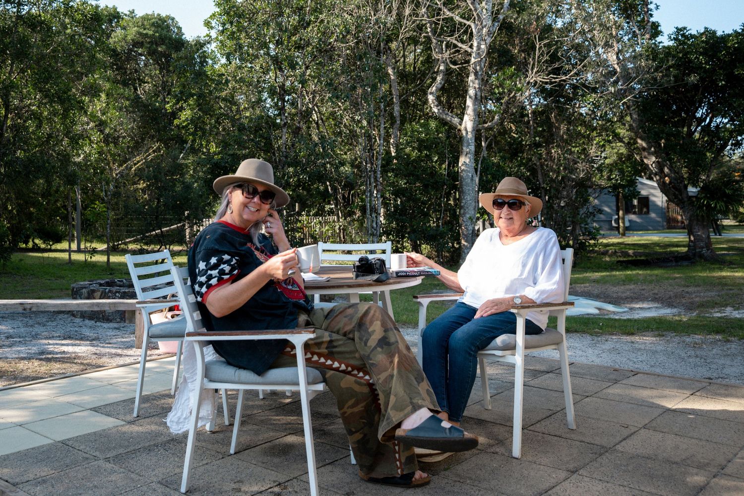 Two people sitting at a white outdoor table enjoying coffee in a sunny park surrounded by green trees.