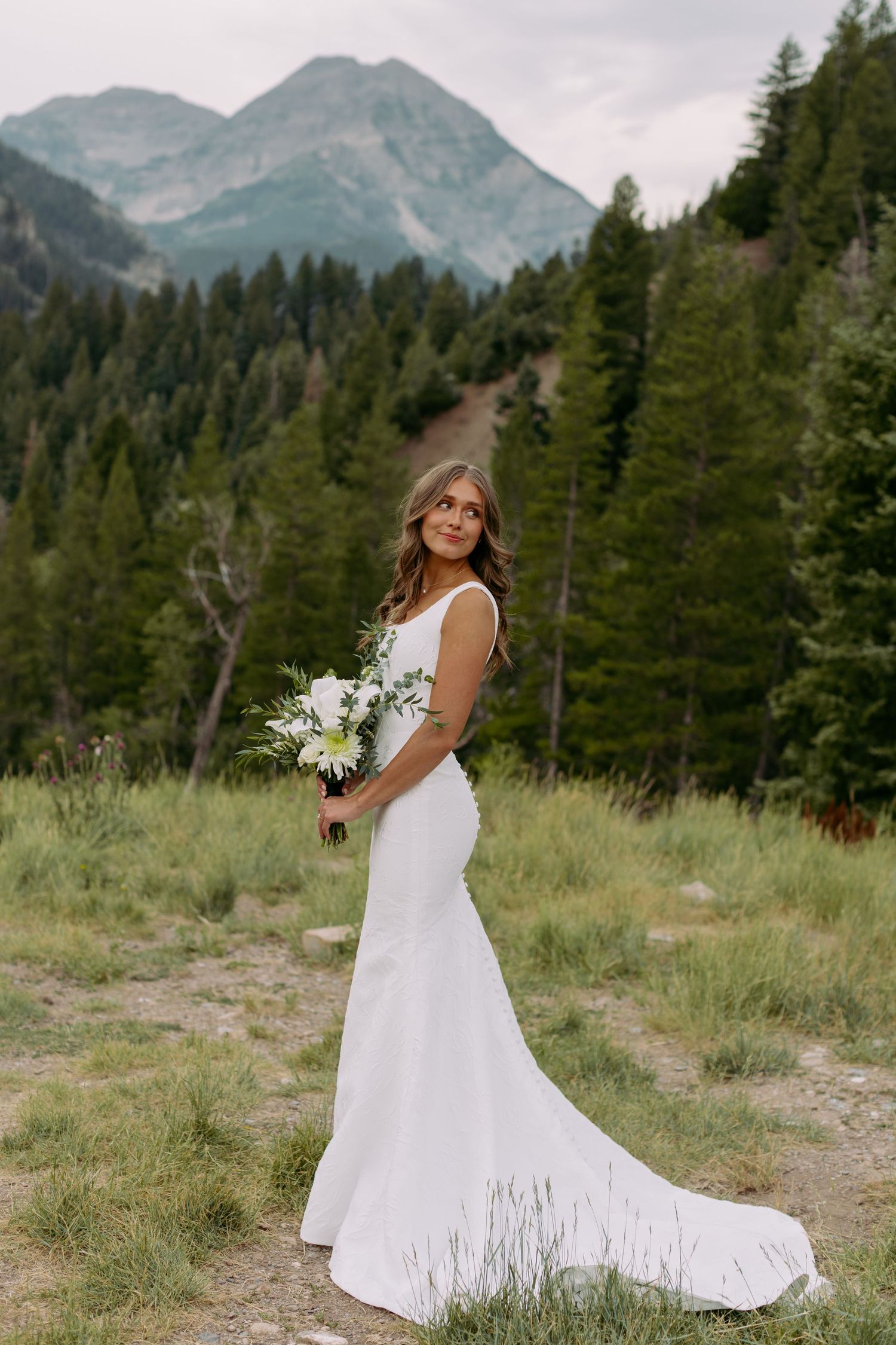 Bride in white mermaid gown holding bouquet poses before mountain forest backdrop.