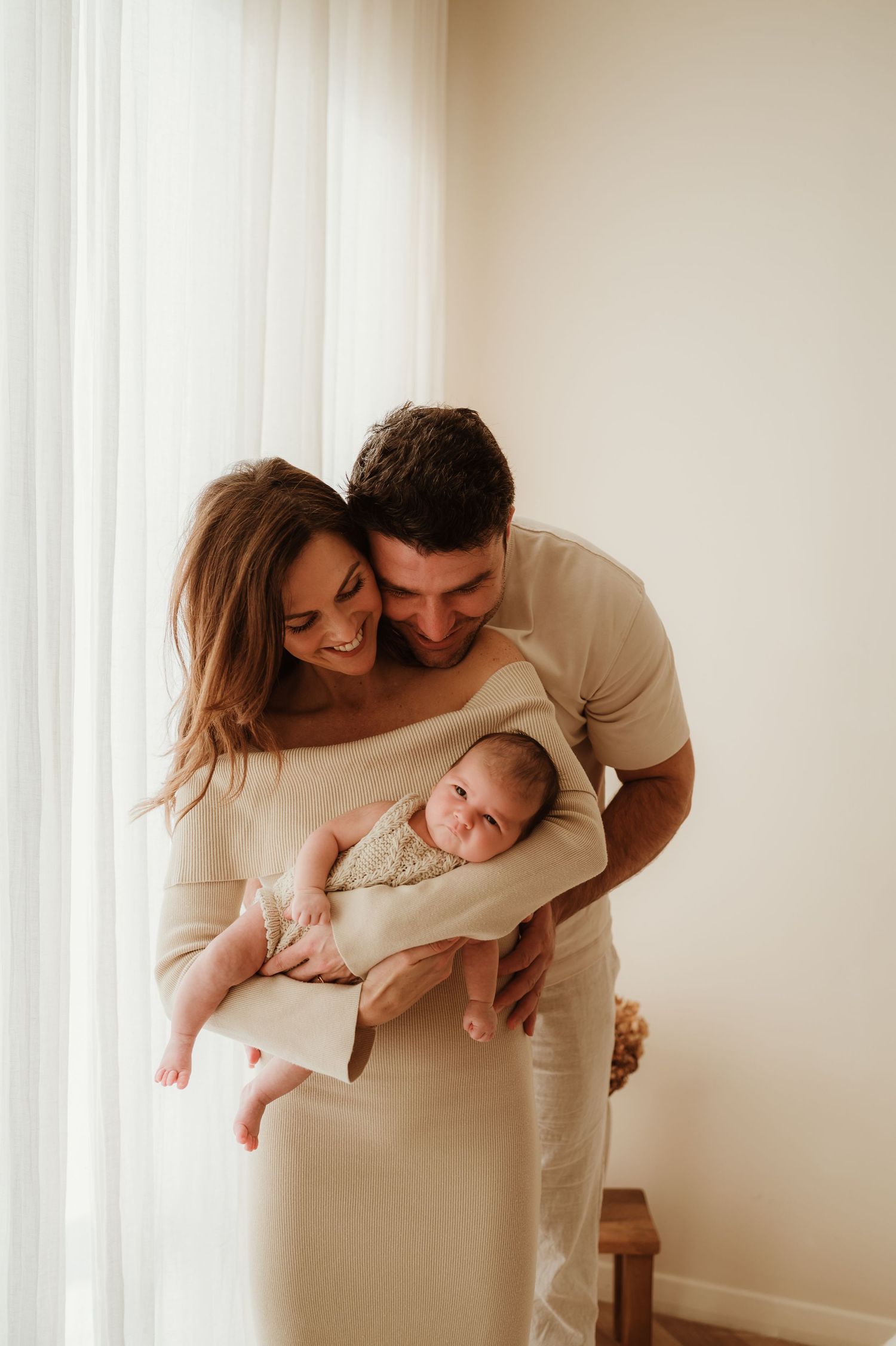 Happy couple in neutral tones holding their swaddled newborn baby by a bright window.