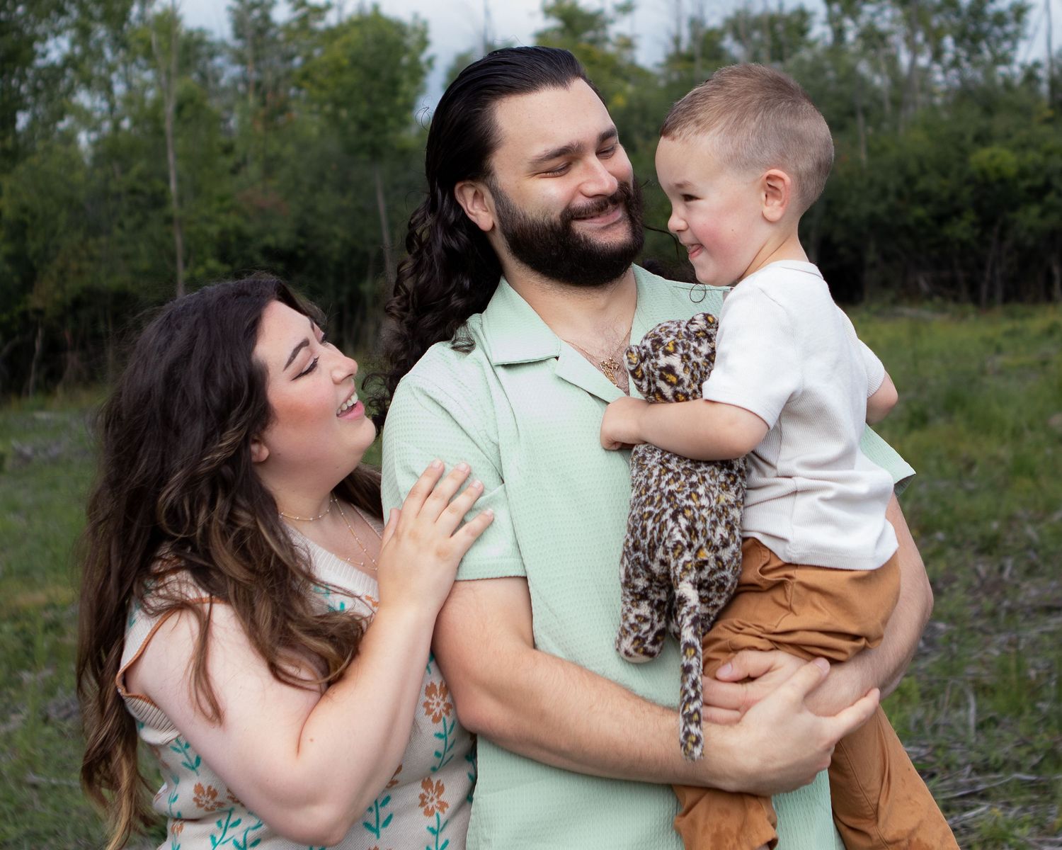 Loving parents holding laughing toddler outdoors with green trees in bright natural background.