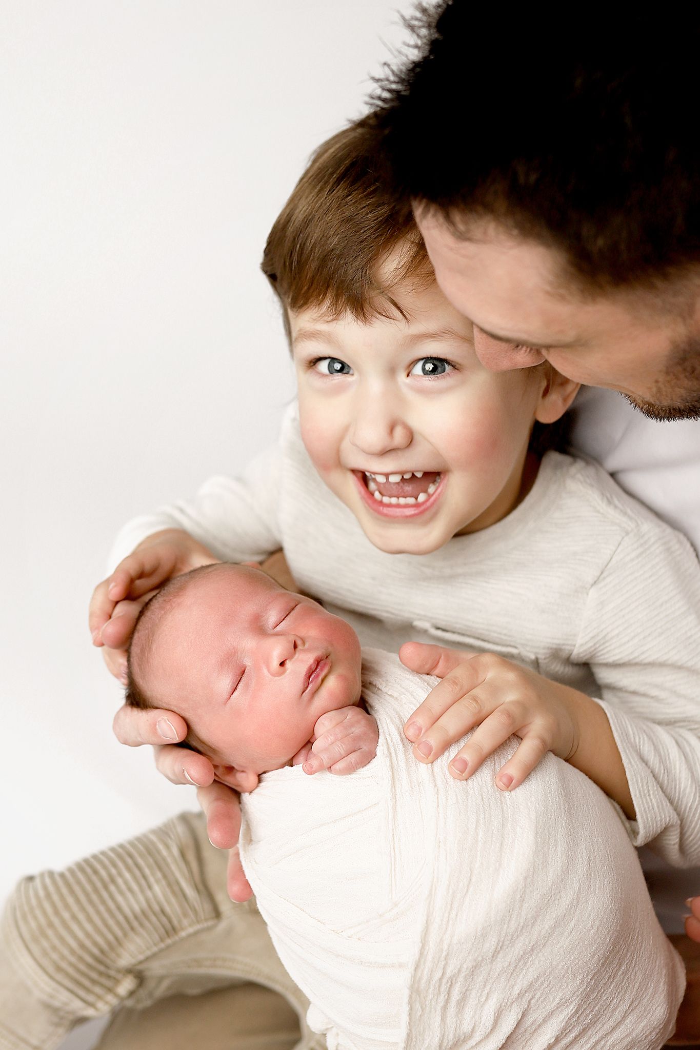 Father holds sleeping newborn while excited toddler smiles broadly, family portrait on white background.