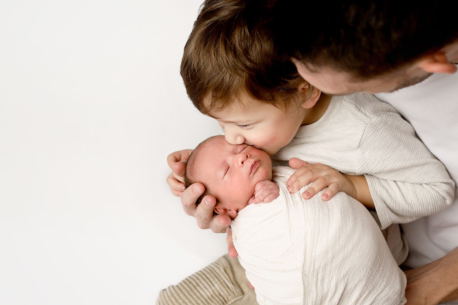 A father and young child tenderly kiss a swaddled newborn baby against a white background. Sibling and newborn photography