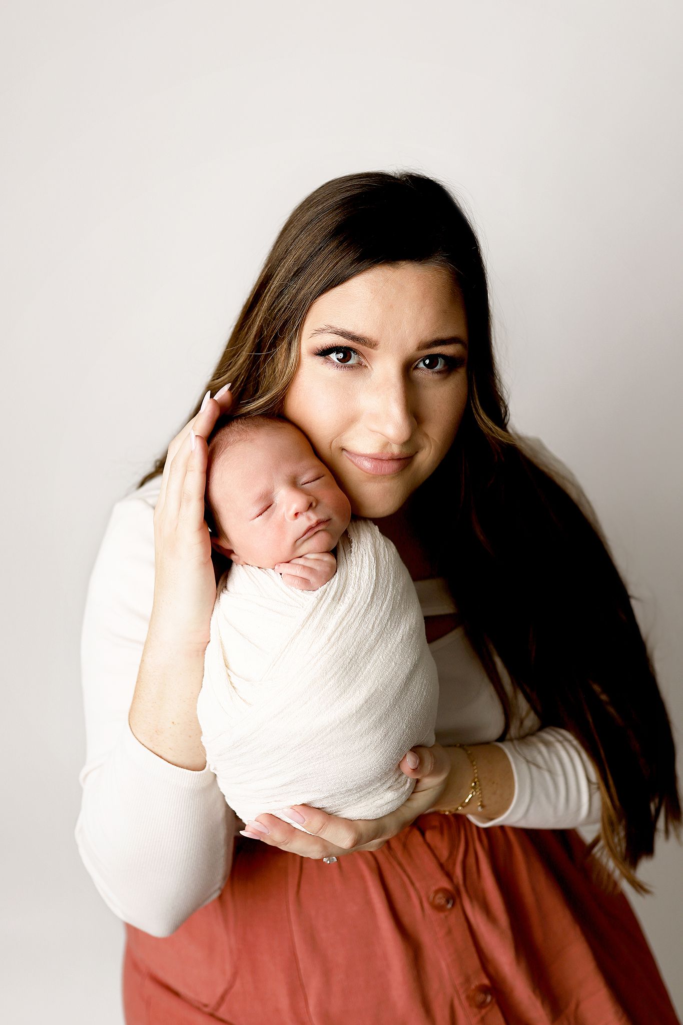 A mother tenderly holds her swaddled newborn baby against her cheek, smiling warmly in a bright studio portrait.