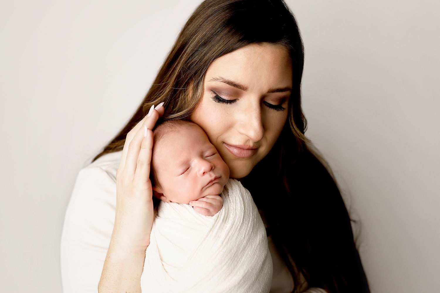 A mother tenderly holds her sleeping newborn baby wrapped in white, against a soft neutral background. Utah newborn photographer