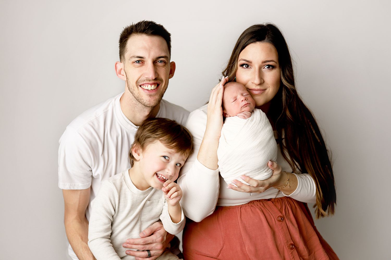 Family of four posing for a newborn portrait, parents smiling with toddler and swaddled baby against a neutral background. Utah photographer