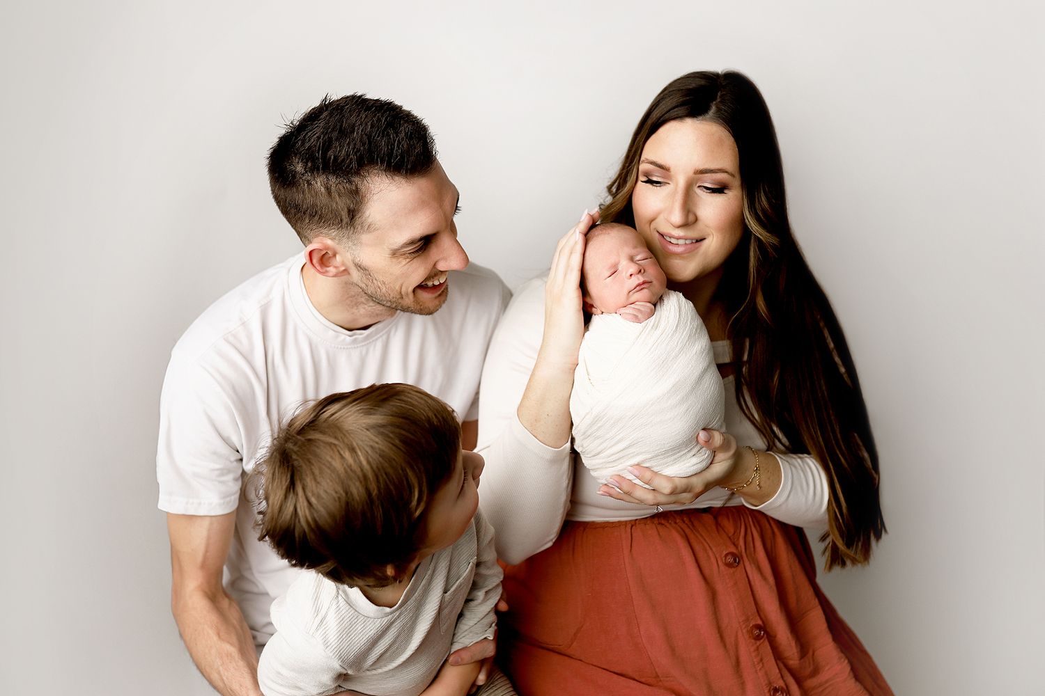 A young family poses together, parents holding a newborn and toddler, smiling warmly against a white background.