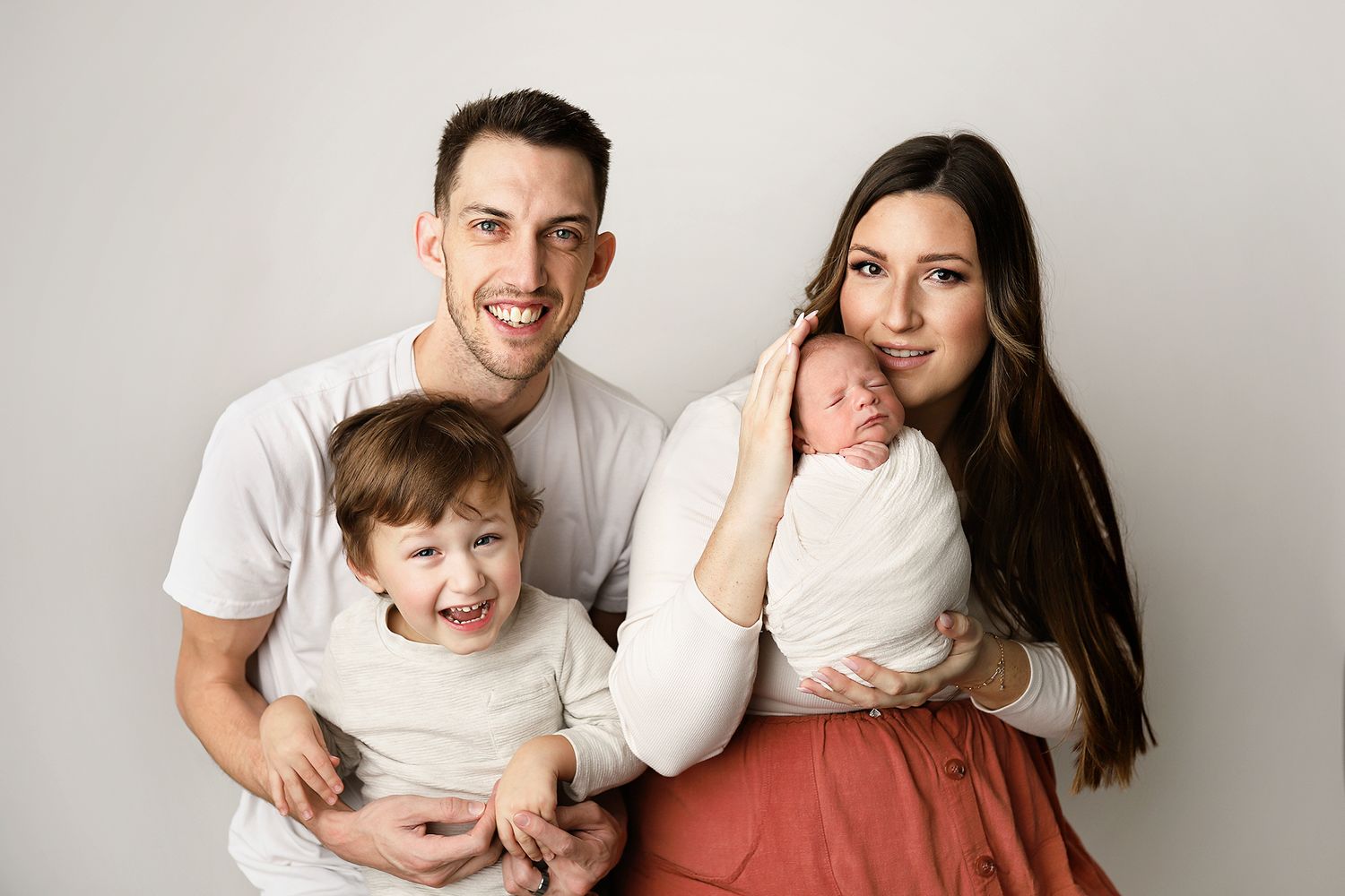 Happy family portrait with parents and two young children, including a newborn baby, posing against a white background.