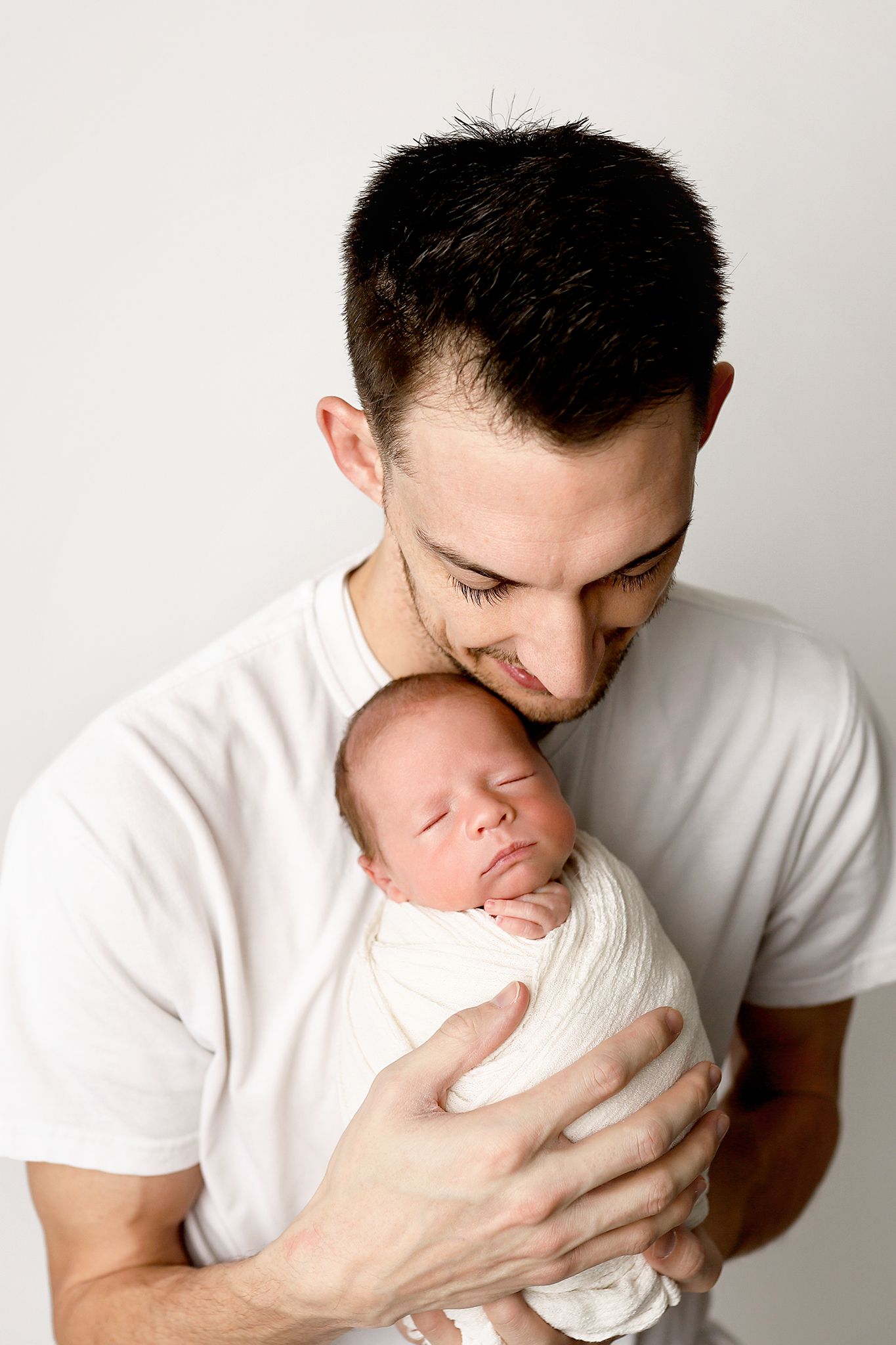 Loving father in white t-shirt gently holding and kissing his sleeping swaddled newborn baby.
