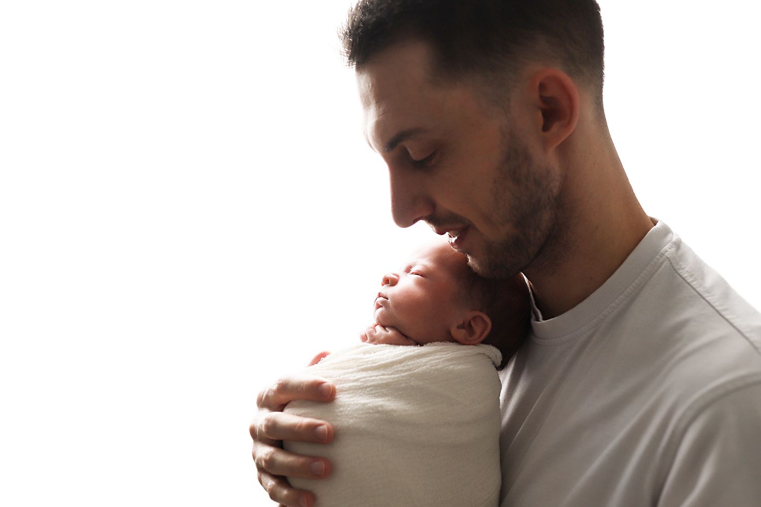 A father tenderly kisses his newborn baby wrapped in a white swaddle against a bright white background. Utah newborn and family photography