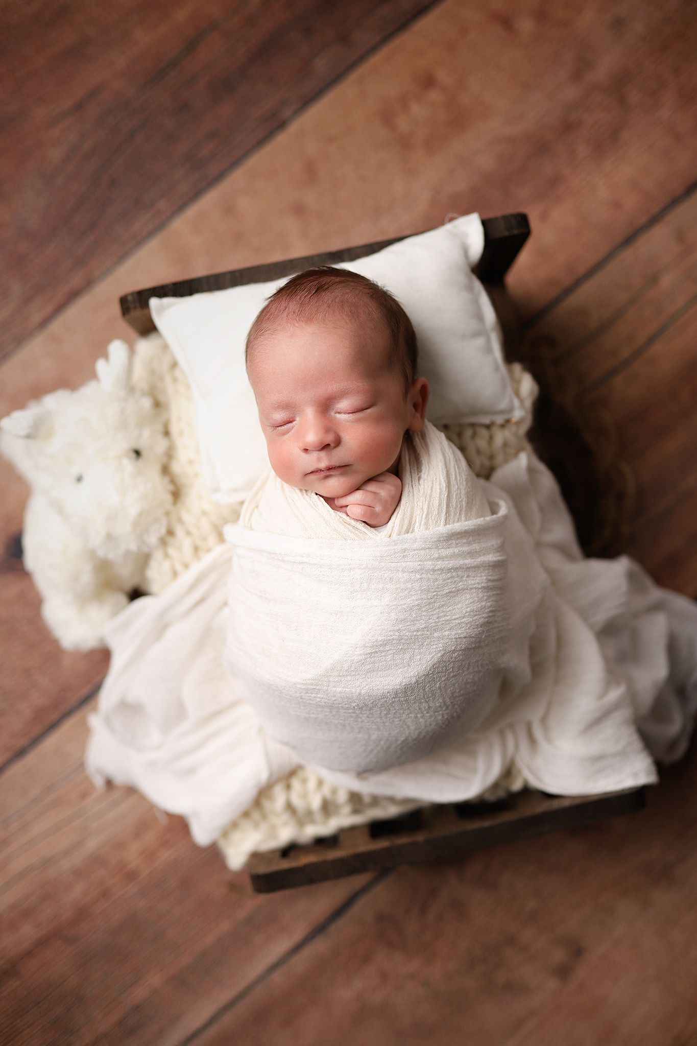 Sleeping newborn baby swaddled in white wrap resting on tiny wooden bed with pillow and stuffed animal. Utah newborn photographer.
