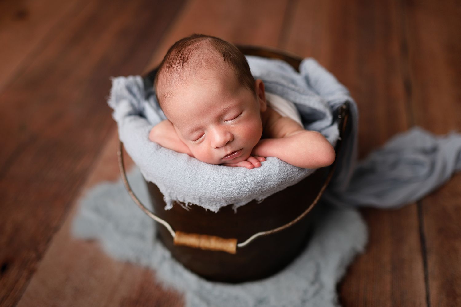 Sleeping newborn baby nestled in a rustic wooden bucket with soft blue fabric on a warm wood floor.