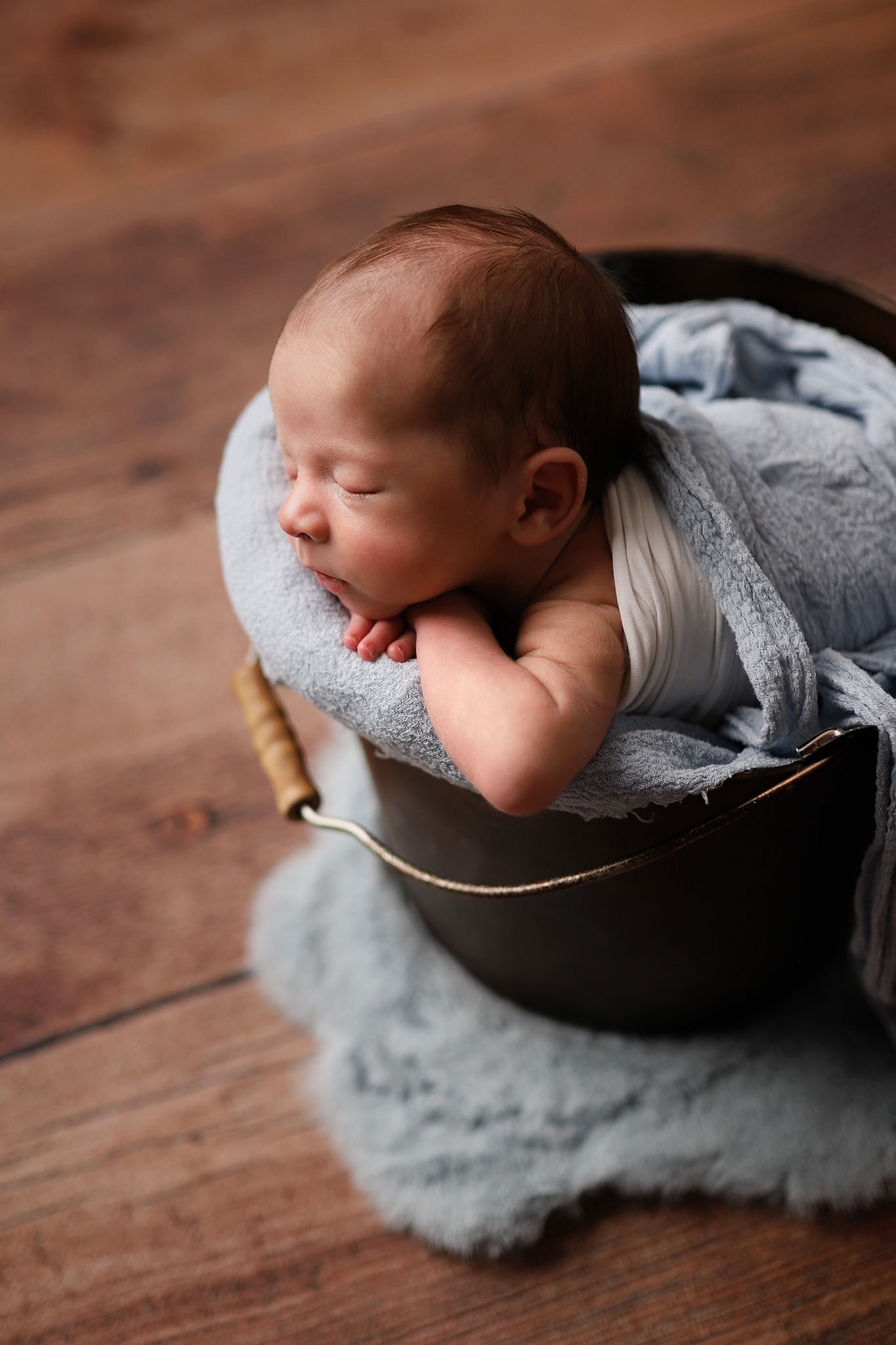 Sleeping newborn baby wrapped in soft blue cloth nestled inside a rustic metal bucket on a wooden floor. Orem newborn photographer