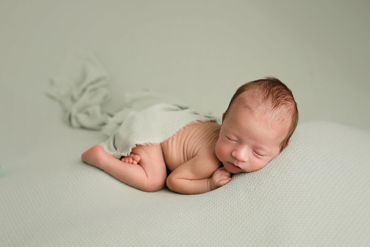 Peaceful newborn baby sleeping on a sage green blanket, curled up in a cozy fetal position during a professional photo session.