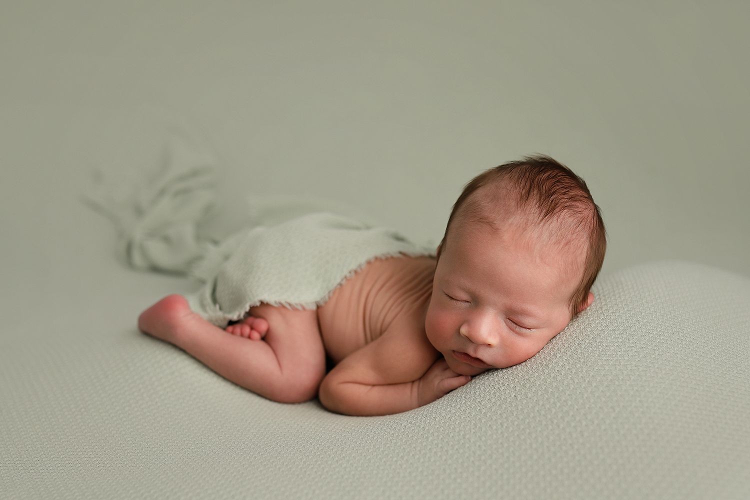 Peaceful newborn baby sleeping on sage green backdrop, wrapped in soft green fabric during professional newborn photography session.