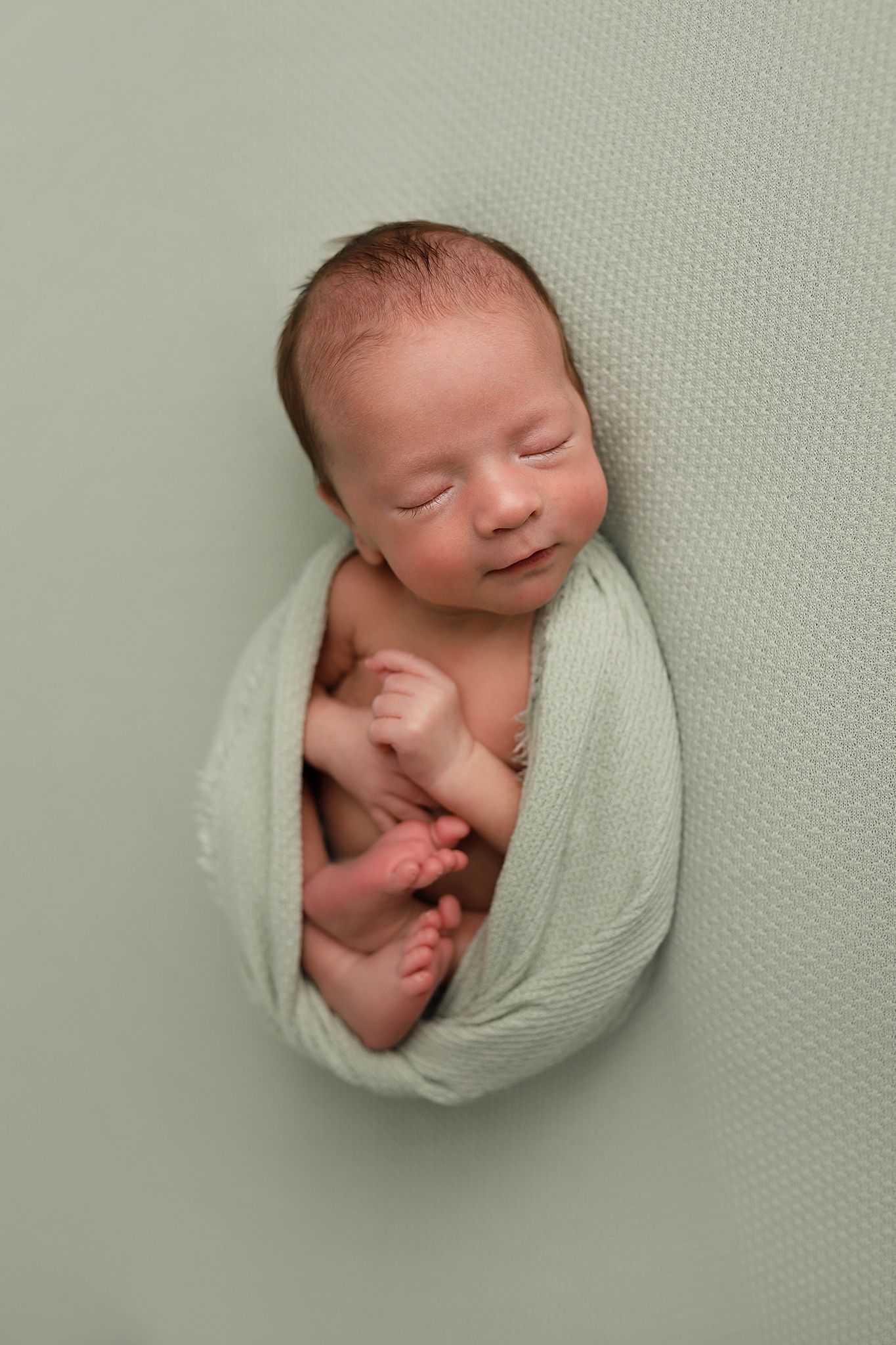 Sleeping newborn baby wrapped in a sage green swaddle blanket with tiny feet peeking out.