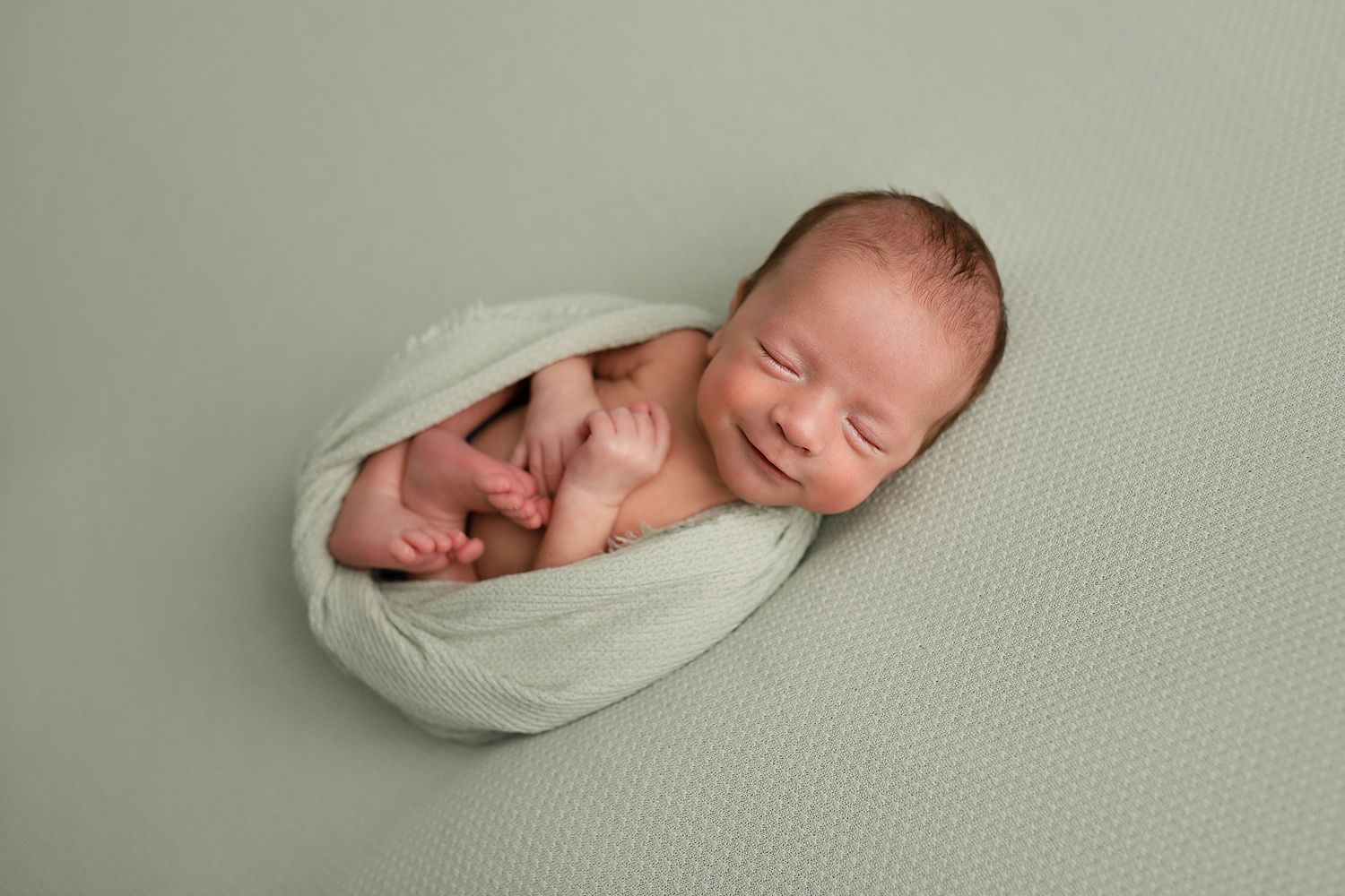 Smiling newborn baby snugly wrapped in a sage green swaddle on a matching green background. Utah newborn photographer